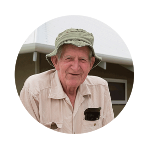 A senior man wearing a green bucket hat and beige shirt smiles at the camera