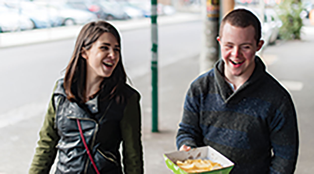 A girl and boy walk down the street laughing. The boy holds takeaway in his hand.