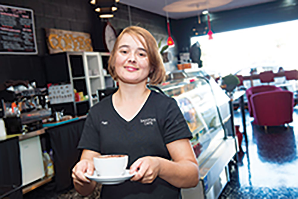 A young waitress at a café walks towards the camera, holding a cup of coffee.